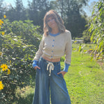 A woman wearing the Contrast Knitted Crew Neck Button Down Cardigan Sweater with denim pants in front of greenery.