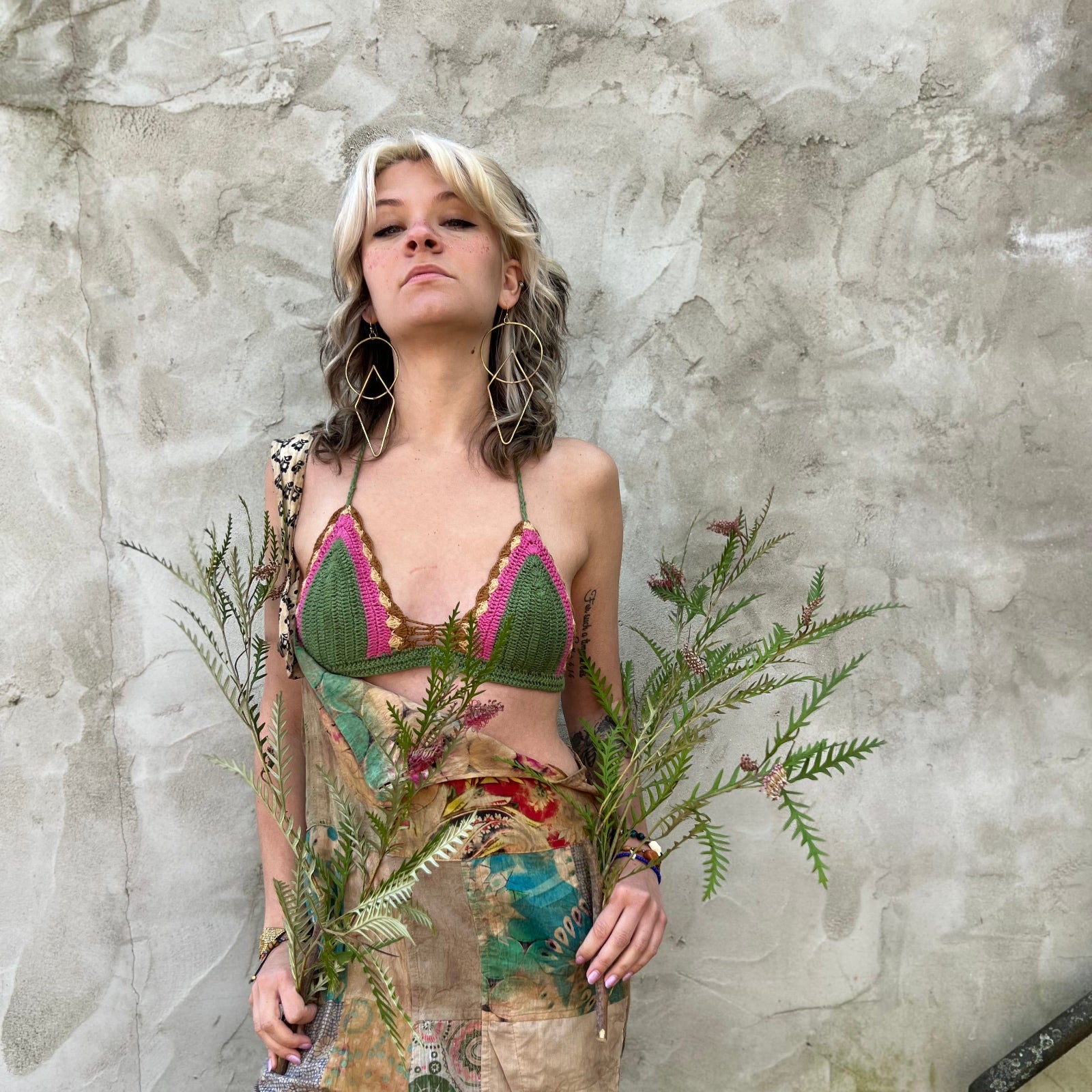 A woman wearing the sage Festive Crochet Top: One Size with patchwork overalls and an evil eye necklace.