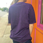 A woman wearing the Purple Mushroom Stonewashed Cotton T-shirt with jeans in front of a pink and orange wall.