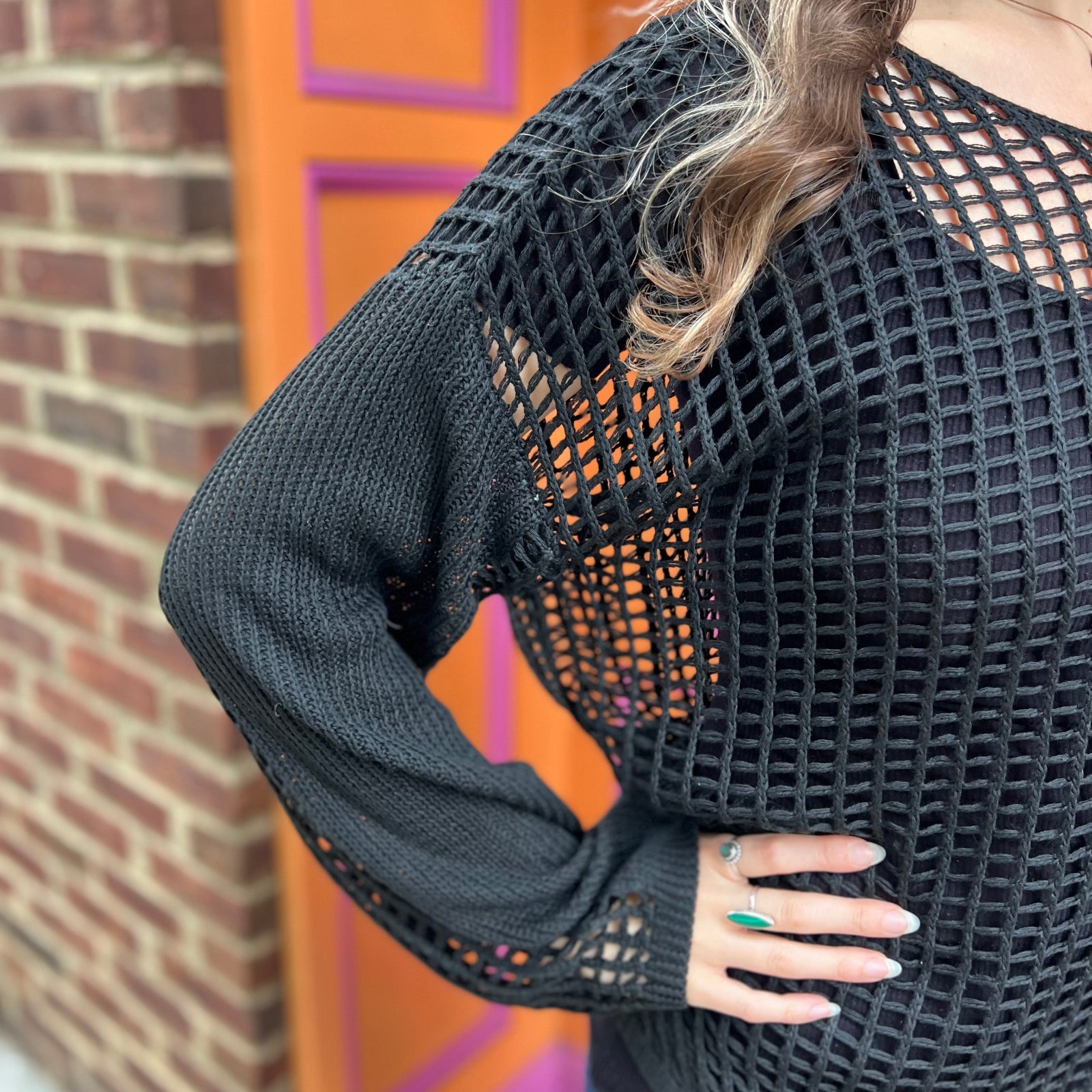 A woman wearing the black Hollow Out Knit Drop Sleeve Sweater in front of a pink and orange wall.