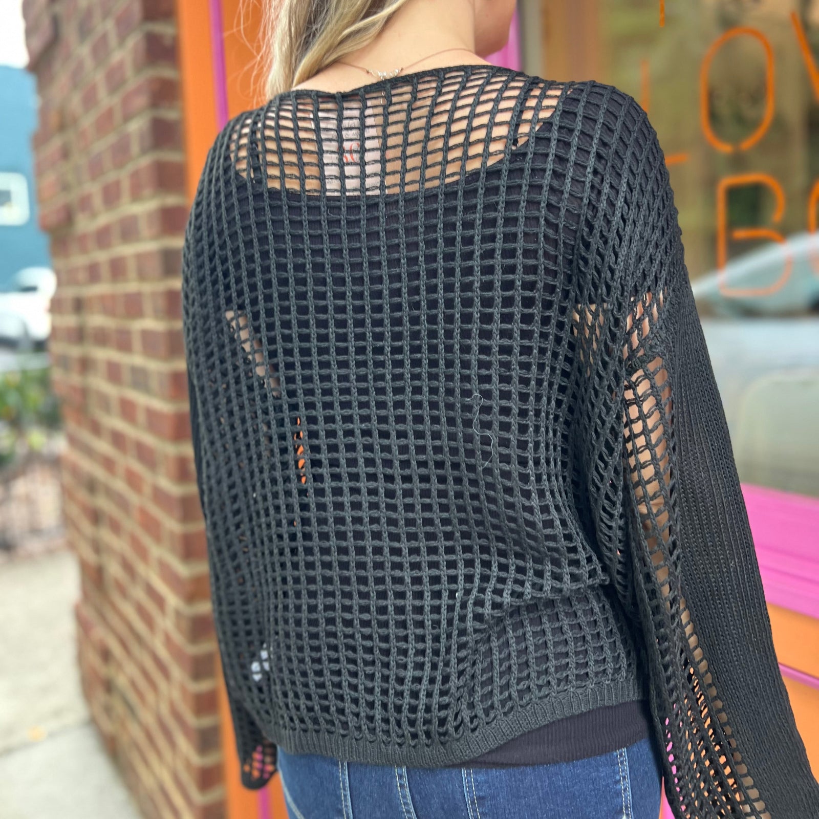 A woman wearing the black Hollow Out Knit Drop Sleeve Sweater in front of a pink and orange wall.