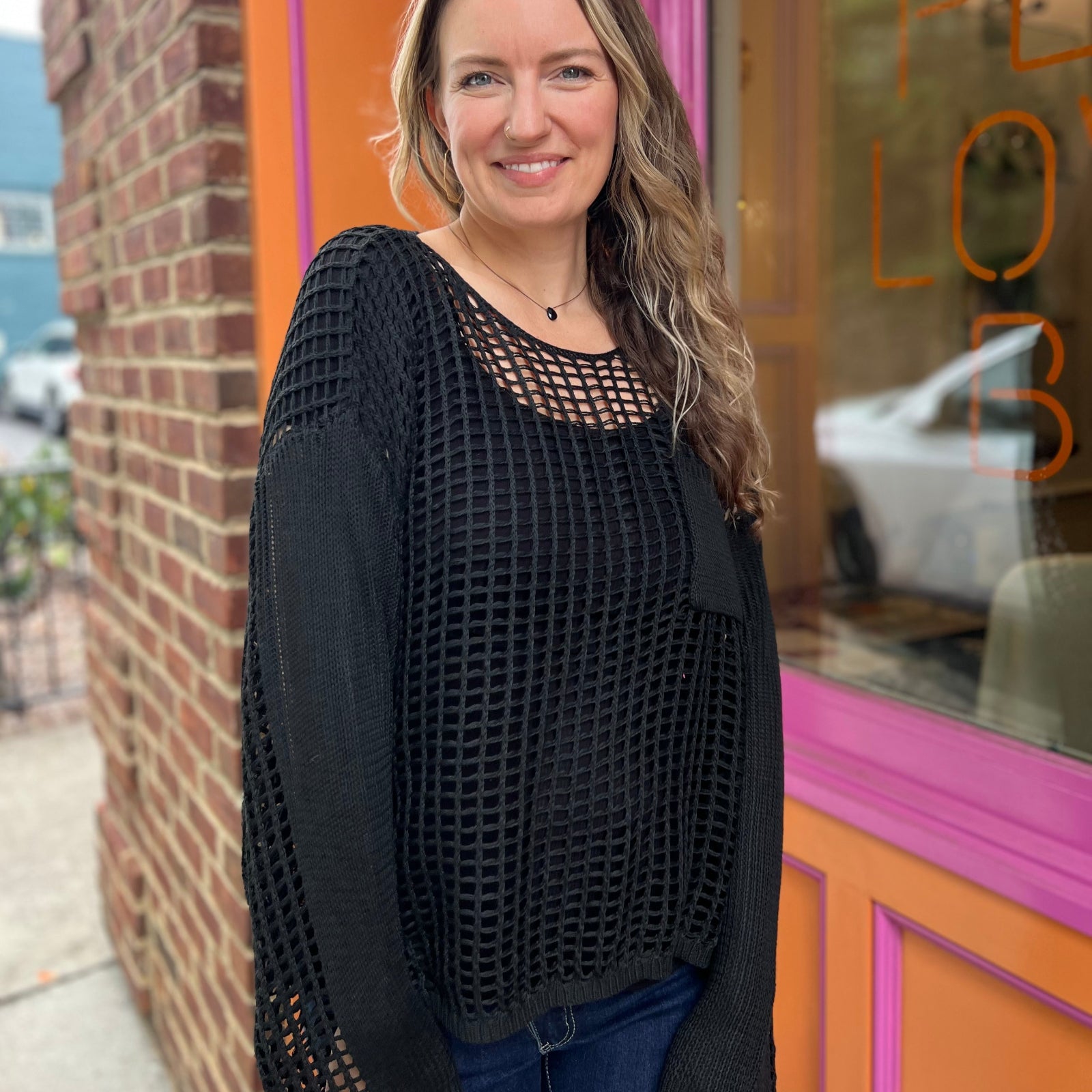 A woman wearing the black Hollow Out Knit Drop Sleeve Sweater in front of a pink and orange wall.