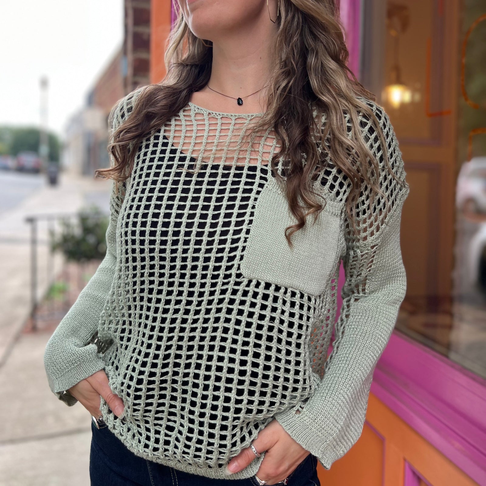 A woman wearing the green Hollow Out Knit Drop Sleeve Sweater in front of a pink and orange wall.