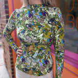 A woman wearing the green Cosmic Mythology Long Sleeve Top with beige linen pants in front of a window.