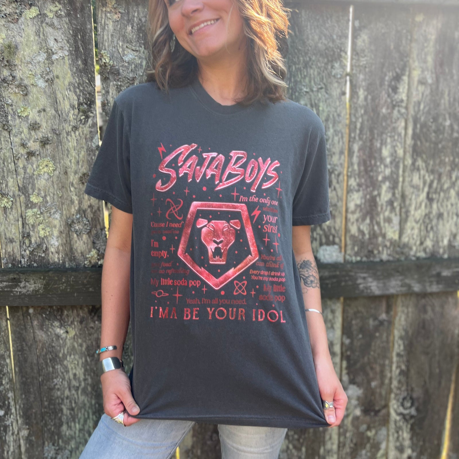 A woman wearing the Saja Boys Kpop Demon Hunters Inspired Cotton Tee with jeans in front of a wooden fence.