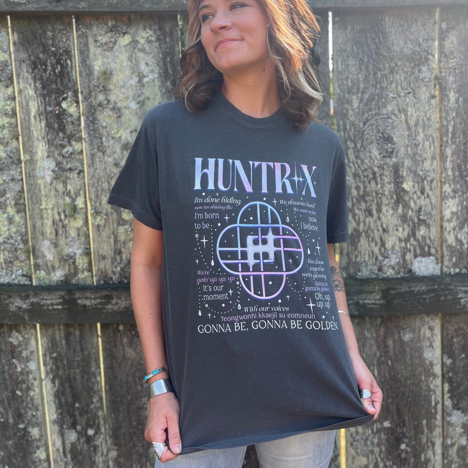 A woman wearing the Huntrix Kpop Demon Hunters Inspired Cotton Tee with jeans in front of a wooden fence.