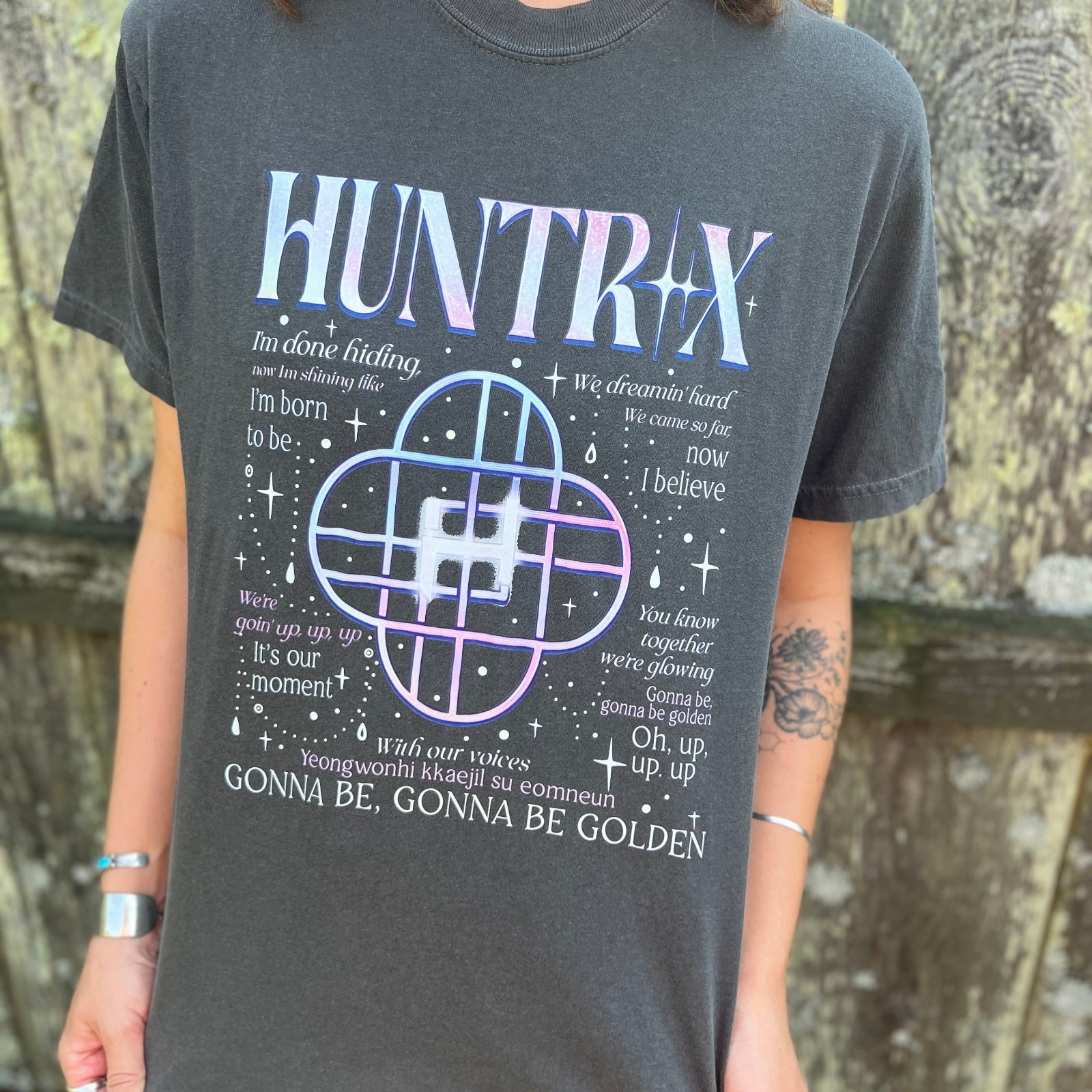 A woman wearing the Huntrix Kpop Demon Hunters Inspired Cotton Tee with jeans in front of a wooden fence.