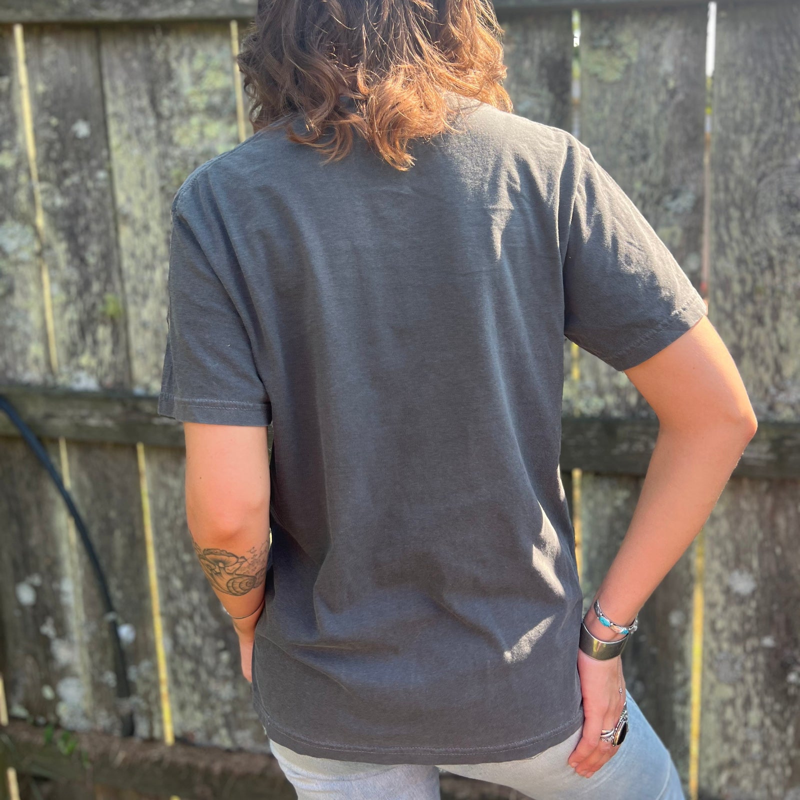 A woman wearing the Huntrix Kpop Demon Hunters Inspired Cotton Tee with jeans in front of a wooden fence, showing off the back.