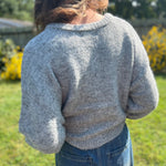 A woman wearing the heather gray Knit Round Neck Sweater Cardigan Top with jeans in front of some greenery.