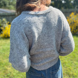 A woman wearing the heather gray Knit Round Neck Sweater Cardigan Top with jeans in front of some greenery.