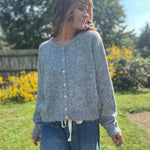 A woman wearing the heather gray Knit Round Neck Sweater Cardigan Top with jeans in front of some greenery.