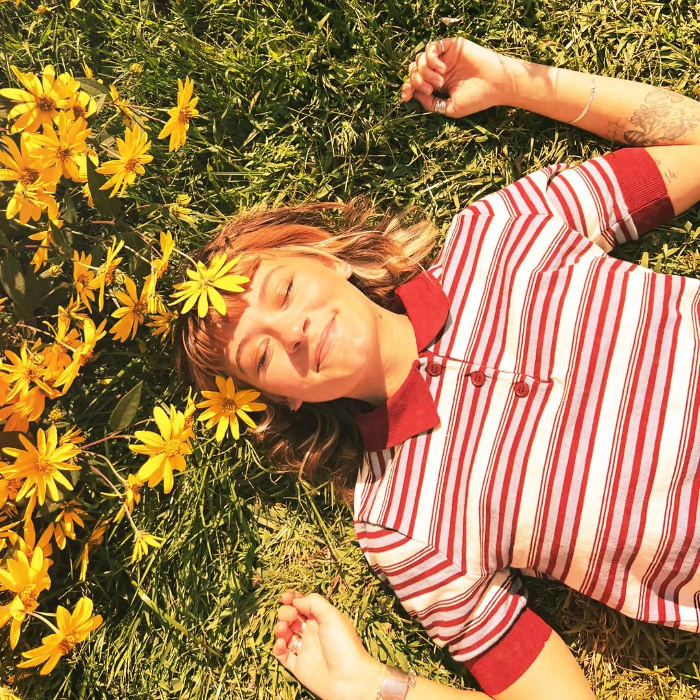 Person lying on grass with yellow flowers around, wearing a red and white retro striped shirt.