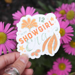 Hand holding a '12 Showgirl' sticker with pink flowers in the background