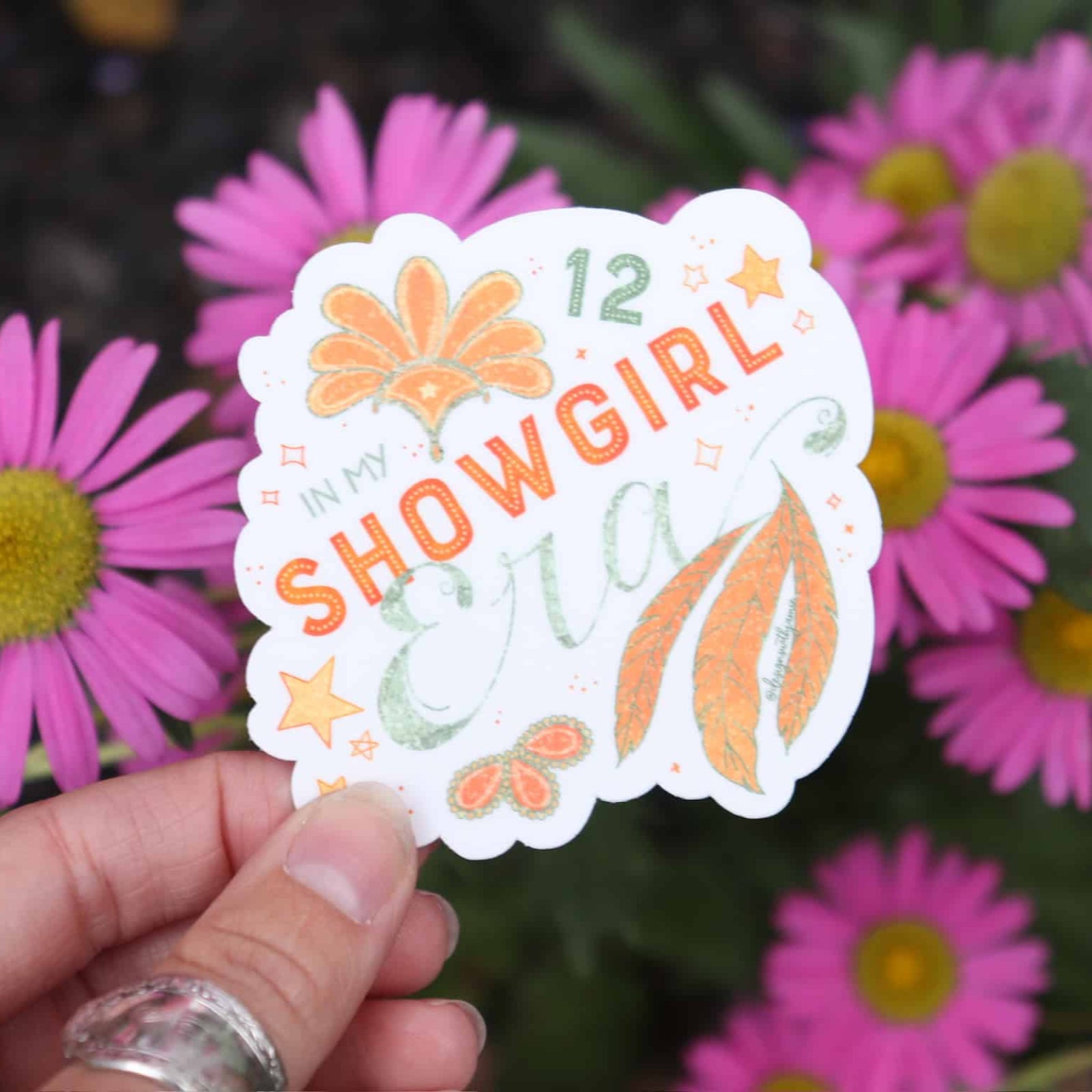 Hand holding a '12 Showgirl' sticker with pink flowers in the background