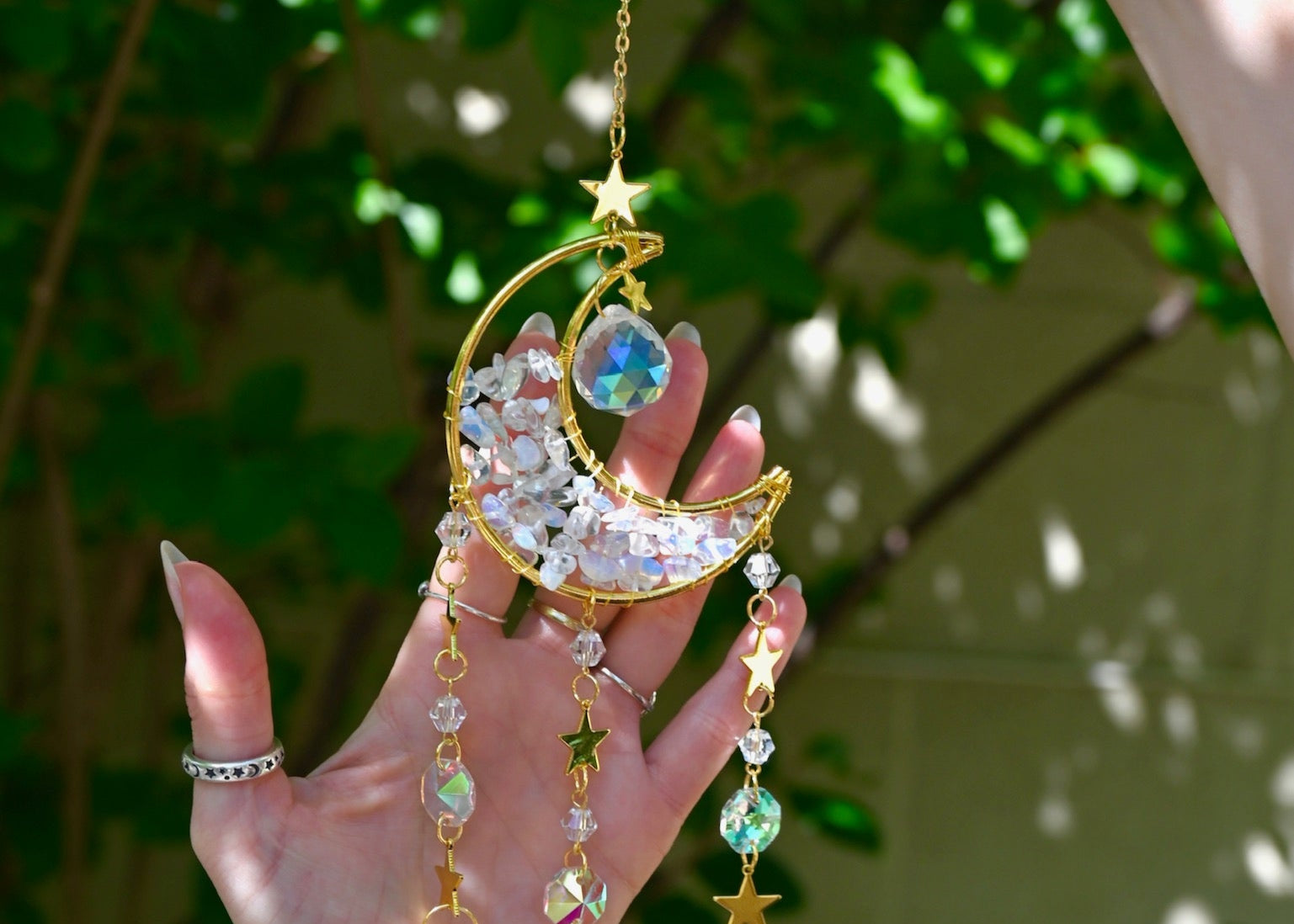 Someone holding the Opalite Crystal Moon Suncatcher with greenery in the background.