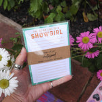 Hand holding a notepad titled 'The To-Do List of a Showgirl' with flowers in the background