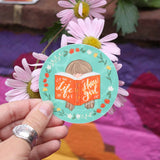 Hand holding a sticker with a design of a girl reading a book, surrounded by flowers, on a colorful fabric background.