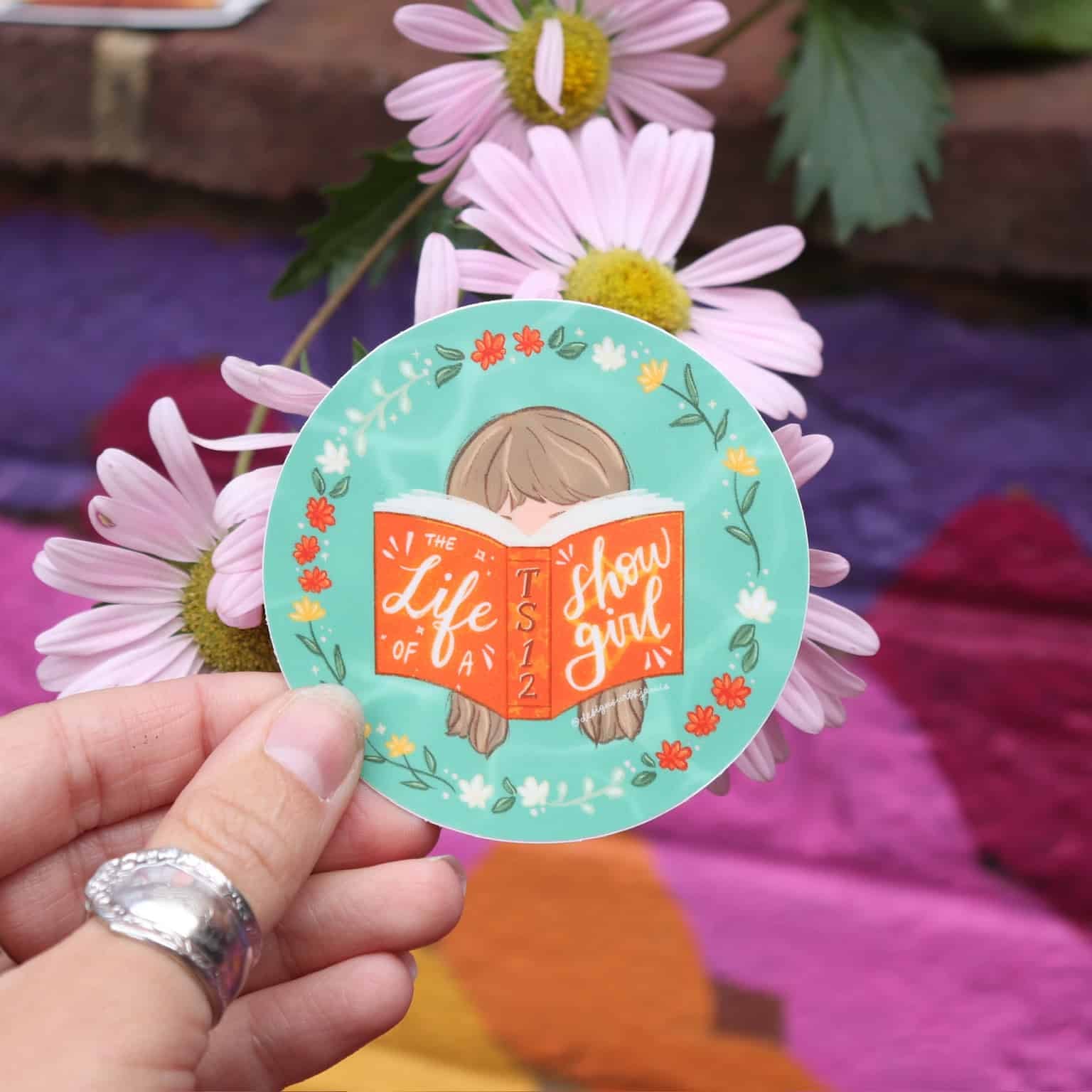 Hand holding a sticker with a design of a girl reading a book, surrounded by flowers, on a colorful fabric background.