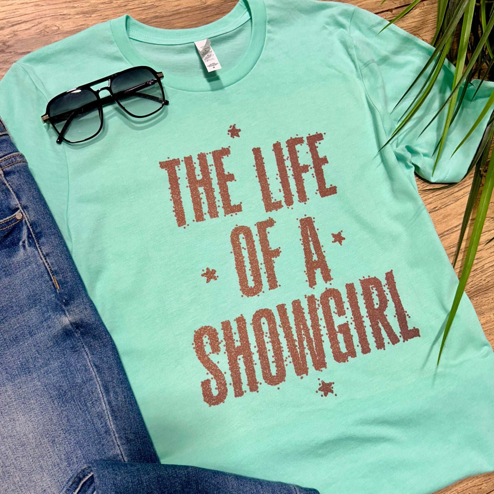 Mint green t-shirt with 'The Life of a Showgirl' text, sunglasses, and jeans on a wooden surface.