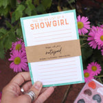 Notepad titled 'The To-Do List of a Showgirl' held by a hand with flowers in the background