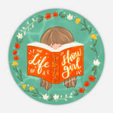 Circular design with a person reading a book titled 'The Life of a TS12 Show Girl' against a green background with flowers.