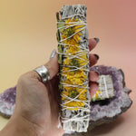 Someone holding the Organic Sunflower White Sage  - 4 in. Smudge Stick in front of an amethyst piece all on a pink and yellow background.