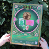 Green and pink 'Dicked' product packaging held by hands against a leafy green background