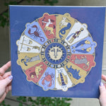 The Mystical Zodiac Print being held in front of a cream-colored wall with greenery.