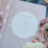 Open book with a pink cover and floral design, surrounded by decorative elements like flowers and stones.