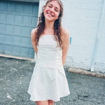 lightweight cream flare dress with square neckline and spaghetti straps in front of white brick