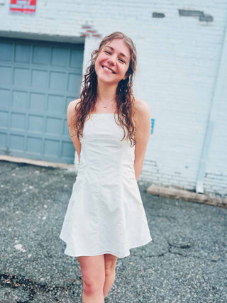 lightweight cream flare dress with square neckline and spaghetti straps in front of white brick