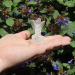 Hand holding a small clear quartz crystal fairy figurine against a background of green foliage and purple flowers