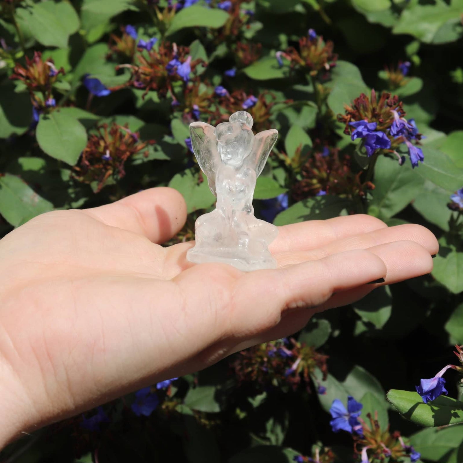 Hand holding a small clear quartz crystal fairy figurine against a background of green foliage and purple flowers