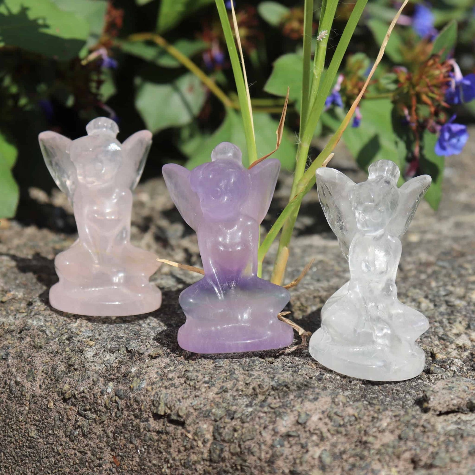 Three fairy figurines made of different colored crystals on a natural stone surface with greenery in the background.