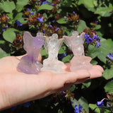 Three crystal fairy crystal figurines held in a hand with a natural background of green leaves and purple flowers.
