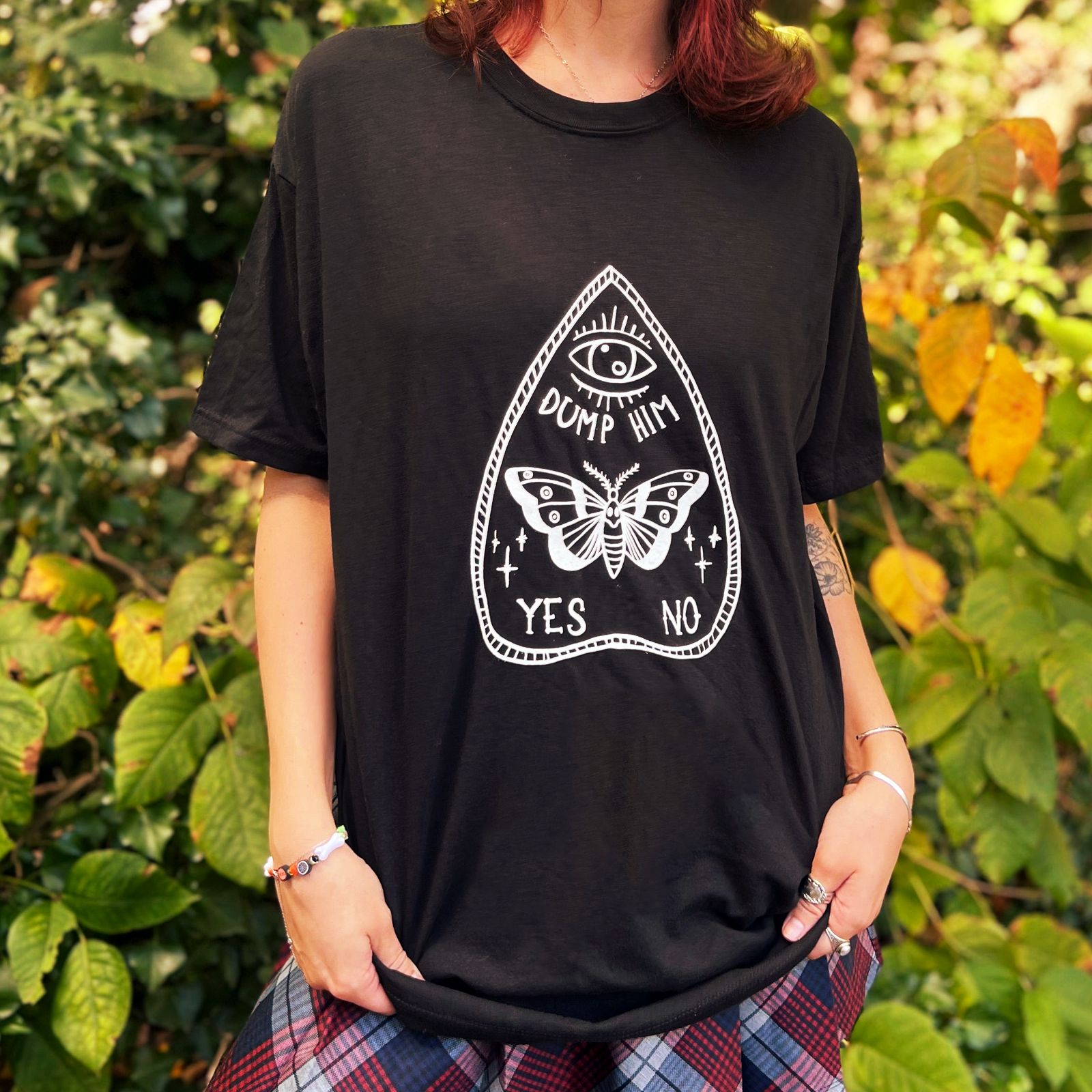 A woman wearing the Dump Him Ouija Cotton T-Shirt with a navy and red plaid skirt in front of greenery.
