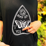 An up-close image of a woman wearing the Dump Him Ouija Cotton T-Shirt and pointing at the shirt, all in front of some greenery.