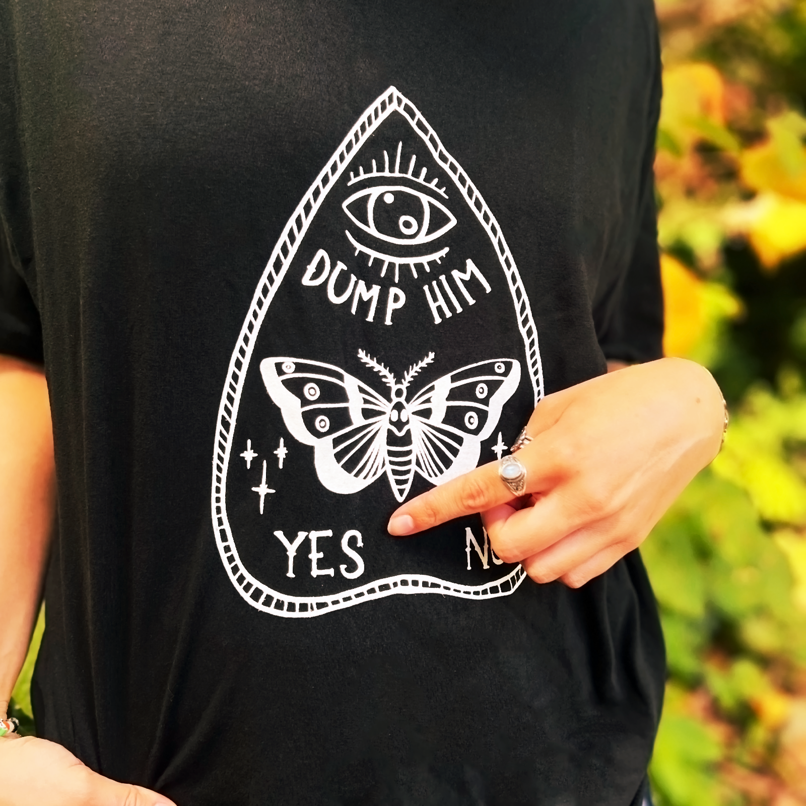 An up-close image of a woman wearing the Dump Him Ouija Cotton T-Shirt and pointing at the shirt, all in front of some greenery.
