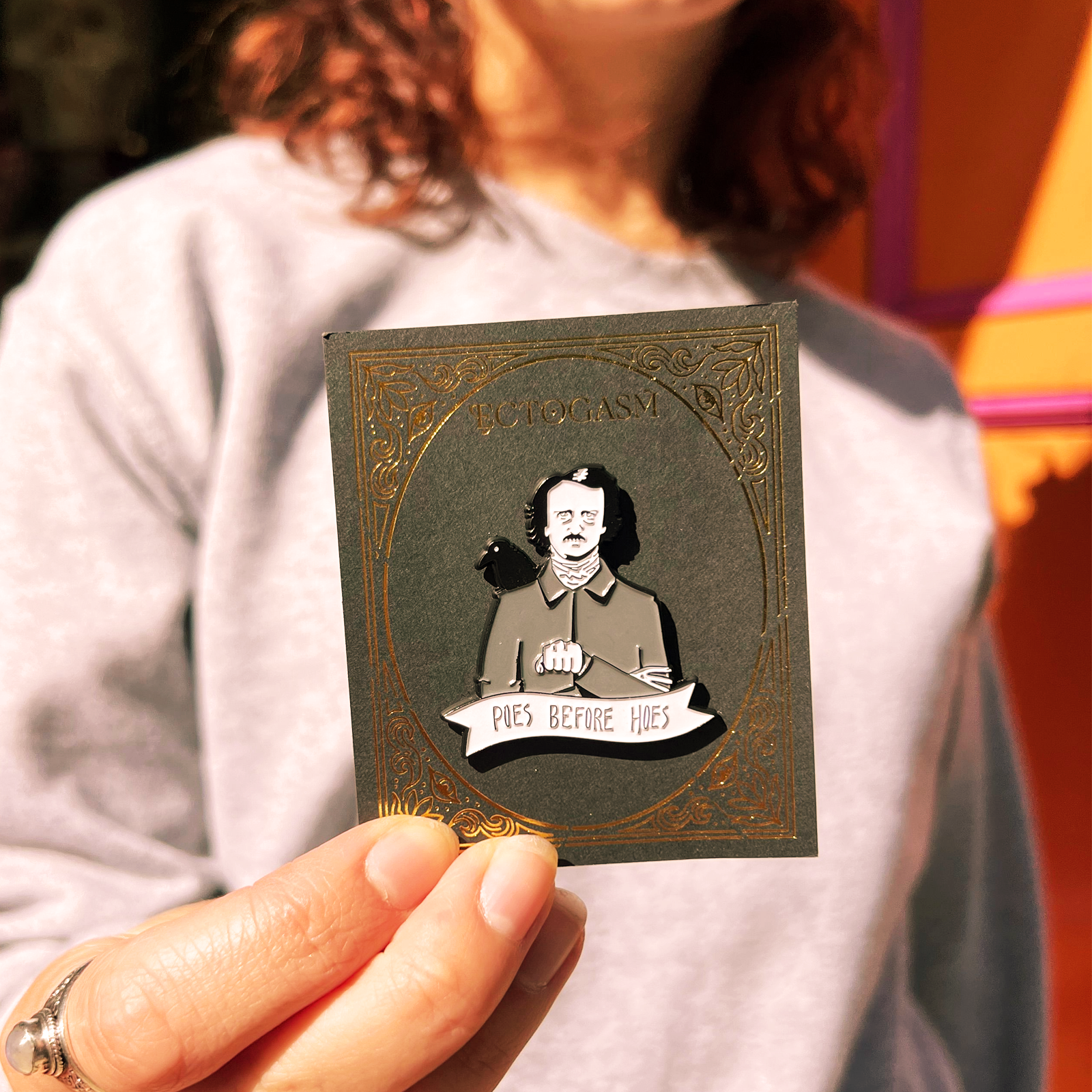 Someone holding the Poes Before Hoes Edgar Allan Poe Enamel Pin in front of themselves.