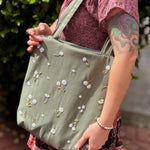 Blonde woman wearing a floral dress and carrying a sage green linen tote bag with hand-embroidered white daisies