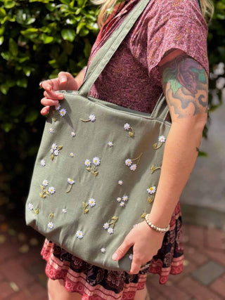 Blonde woman wearing a floral dress and carrying a sage green linen tote bag with hand-embroidered white daisies