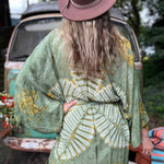 Person wearing a green patterned kimono and brown hat, standing outdoors with a vintage car in the background.