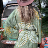 Person wearing a green patterned kimono and brown hat, standing outdoors with a vintage car in the background.
