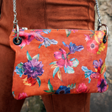 Someone wearing the Orange Floral Print Italian Leather Crossbody Bag with burnt orange velvet pants in front of a stone background.