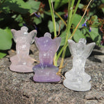 Three crystal carved fairy figurines in different colors on a stone surface with greenery in the background.