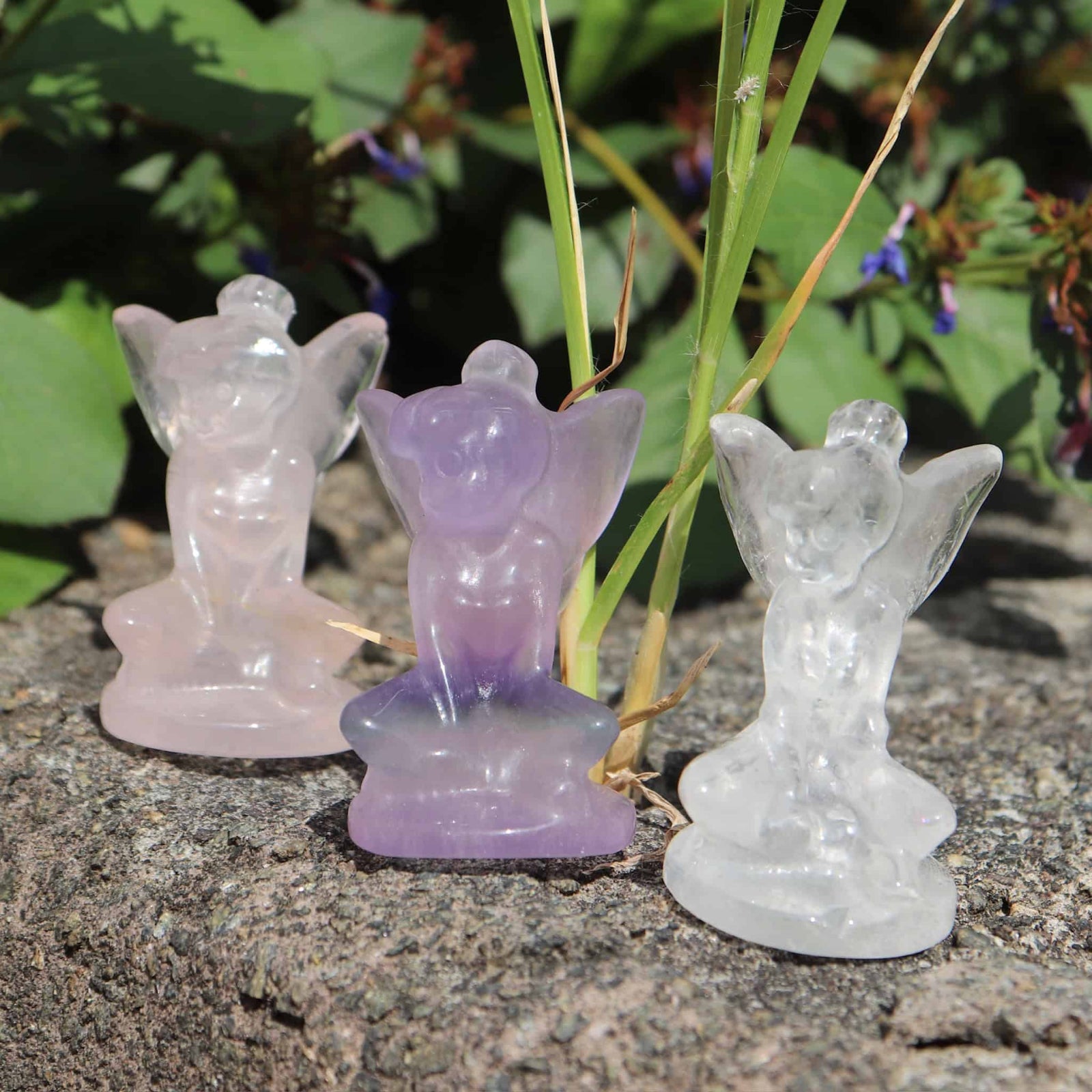 Three crystal carved fairy figurines in different colors on a stone surface with greenery in the background.
