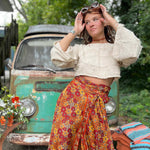 Woman in a white blouse and colorful skirt standing in front of a vintage car with greenery in the background