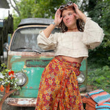 Woman in a white blouse and colorful skirt standing in front of a vintage car with greenery in the background