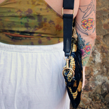 Someone wearing the Small Black and Gold Heron Velvet Crossbody Bag with a white skirt and a tie dye tank.