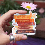Hand holding a decorative sticker with text in front of a blurred floral background